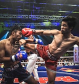 Two athletes are engaged in a dynamic kickboxing match inside a ring, surrounded by a cheering audience. One fighter, wearing red gloves and shorts, executes a high kick towards his opponent, who attempts to block the attack with his arm. The background features colorful lighting and promotional banners.