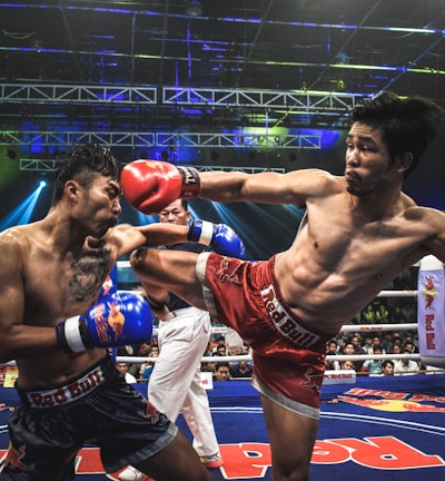 Two athletes are engaged in a dynamic kickboxing match inside a ring, surrounded by a cheering audience. One fighter, wearing red gloves and shorts, executes a high kick towards his opponent, who attempts to block the attack with his arm. The background features colorful lighting and promotional banners.