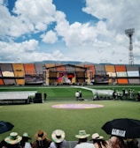 A sports stadium with a large stage set up on the field, hosting an outdoor event. Spectators wearing colorful hats sit in the foreground. The seating areas display vibrant colors, and a clear blue sky with scattered clouds is visible. Large screens display graphics related to the event.
