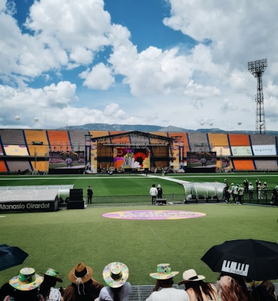 A sports stadium with a large stage set up on the field, hosting an outdoor event. Spectators wearing colorful hats sit in the foreground. The seating areas display vibrant colors, and a clear blue sky with scattered clouds is visible. Large screens display graphics related to the event.