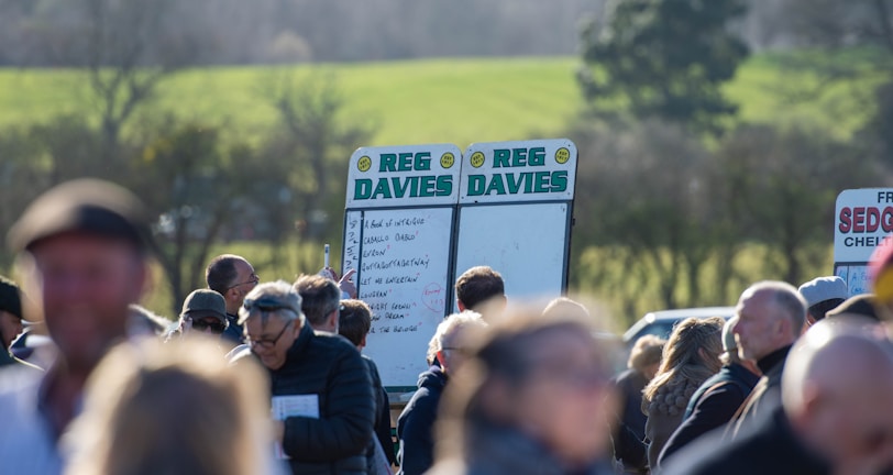 A group of people gathered outdoors with two large betting boards in the background labeled 'REG DAVIES'. The setting appears to be a countryside location with trees and fields visible in the distance. The crowd consists of individuals dressed in jackets and hats, engaging in conversation.