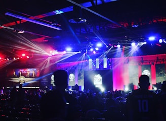 A dimly lit indoor arena filled with an audience. Bright stage lights in red and blue illuminate multiple large screens displaying graphics.