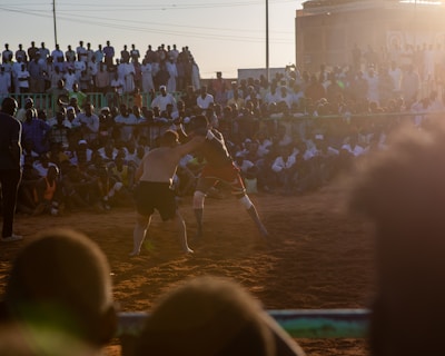 A vibrant wrestling match taking place outdoors, surrounded by a large crowd of spectators. The atmosphere is charged with excitement as people stand and sit around the dirt arena, intently watching the competitors. The sun is setting, casting a warm glow over the scene.