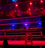 An empty boxing ring is illuminated by dramatic red and blue spotlights inside a dimly-lit arena. A small stool is placed in the center of the ring, surrounded by ropes. The arena is adorned with banners depicting action scenes, and a few silhouettes of spectators can be seen in the background.