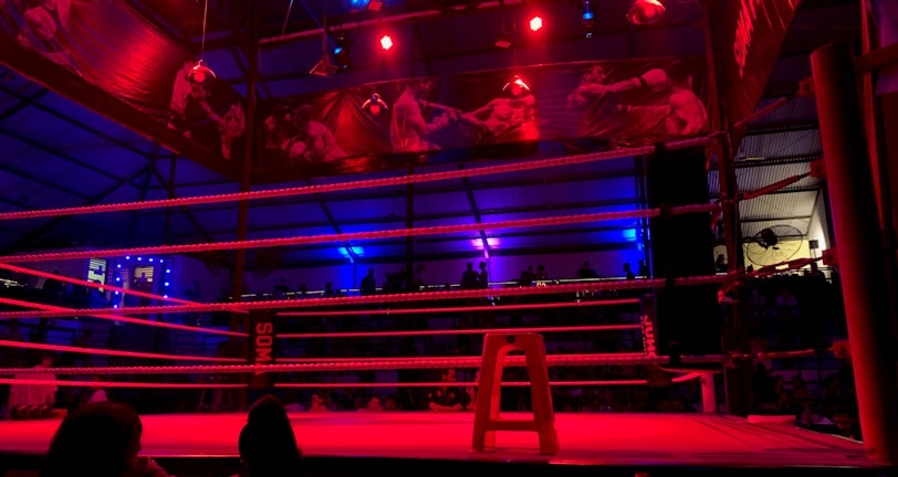 An empty boxing ring is illuminated by dramatic red and blue spotlights inside a dimly-lit arena. A small stool is placed in the center of the ring, surrounded by ropes. The arena is adorned with banners depicting action scenes, and a few silhouettes of spectators can be seen in the background.