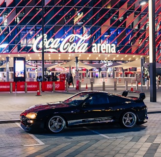 A sleek black sports car is parked in front of an illuminated arena entrance, with a large sign bearing the name of a famous beverage brand. The building features a modern, geometric facade with bright red and blue lights, creating a vibrant and lively atmosphere.