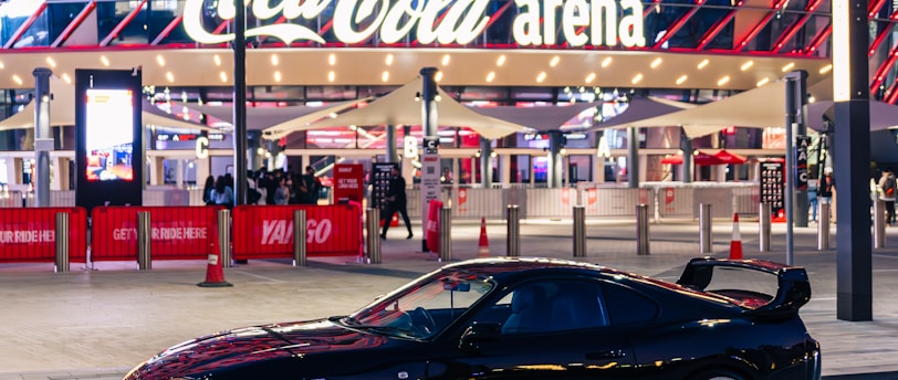 A sleek black sports car is parked in front of an illuminated arena entrance, with a large sign bearing the name of a famous beverage brand. The building features a modern, geometric facade with bright red and blue lights, creating a vibrant and lively atmosphere.